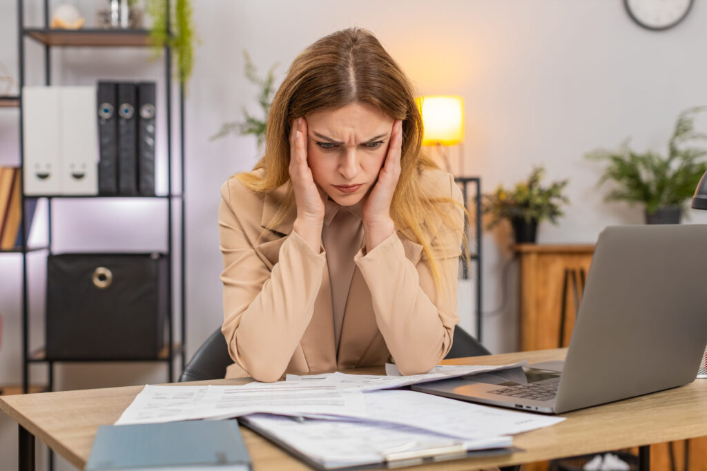 Middle-aged businesswoman at home office looks at bills invoices receipt on laptop sees costs go up frowns confused and unhappy. Freelancer girl at table writes savings plan sets targets prints page