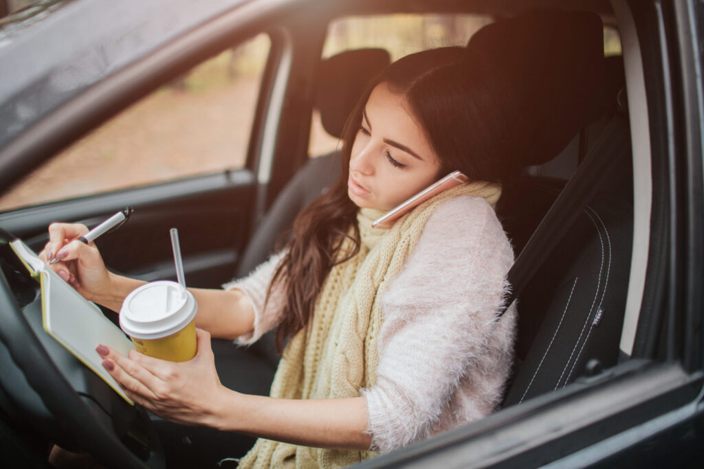 Busy woman is in a hurry, she does not have time, she is going to worck on the go. Worker is drinking hot coffee, writing in a notebook and talking on the phone at the same time. Business female person. She multitasking while using on the phone and driving a car