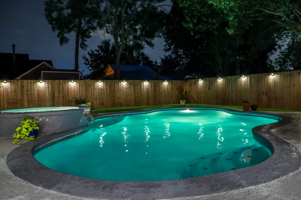 A backyard swimming pool and jacuzzi hot tob at night with solar lights around the fence for privacy and illumination.