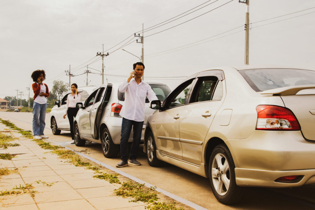Man who was involved in a three vehicle collision standing stressed looking at the damage use phone to call the insurance company to clear and assess the initial damage to the other two women's cars.