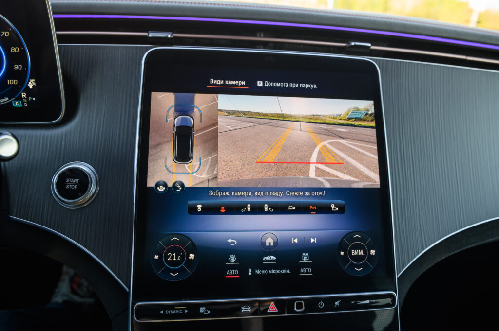 Large touchscreen infotainment display in a luxury car showing a 360-degree backup camera view and parking assist system with active guidelines, close up.