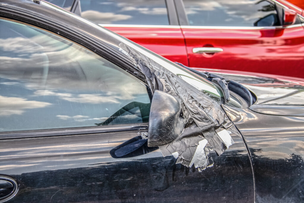 Closeup of dirty car with right side mirror taped on and scratches to it and window - lots of reflections of blue sky and a bright red blurred car in the background